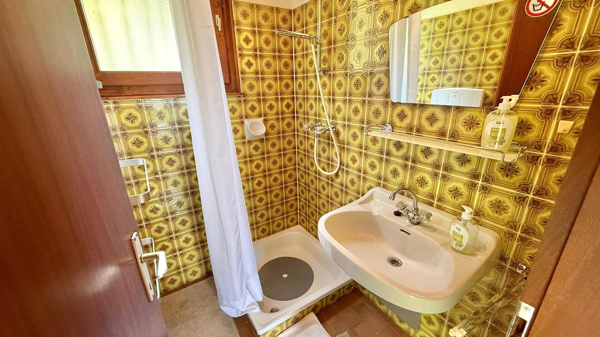 Badezimmer mit gelb-braunen Fliesen, Waschbecken und Duschbereich, im Stil der 80er Jahre.