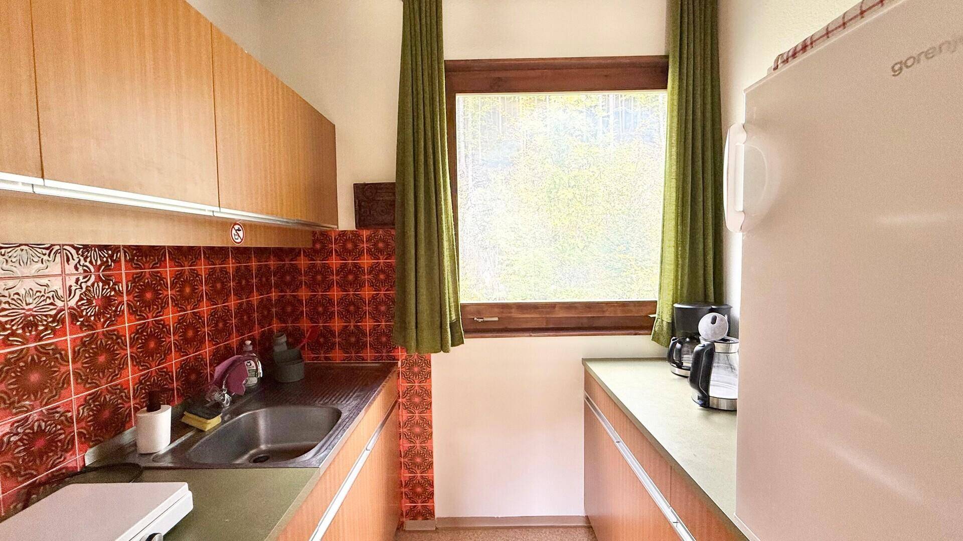 Kleine Küche mit orangefarbenen Fliesen, Holzschränken und einem Fenster mit Blick ins Grüne.