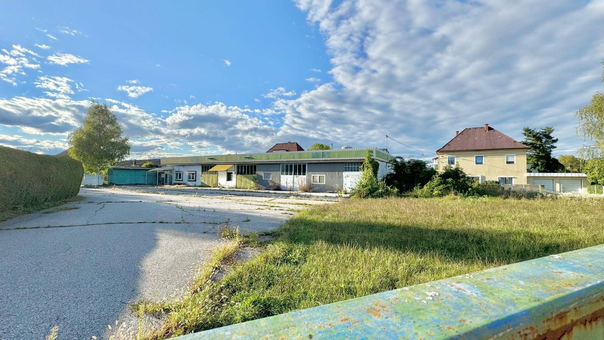 Außenansicht des Gebäudes an einem sonnigen Tag mit angrenzendem Wohnhaus und weitläufiger Grünfläche.