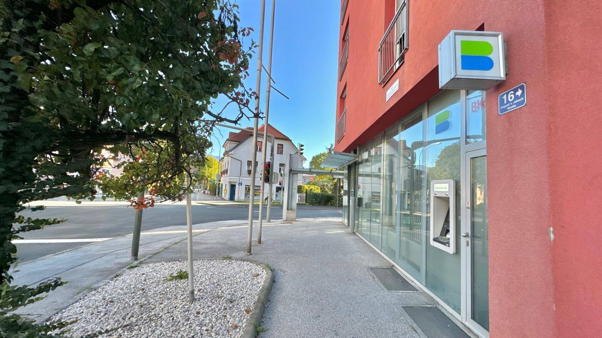 Gebäudefassade mit Bankautomat und Hausnummer an einer sonnigen Straße mit Bäumen.