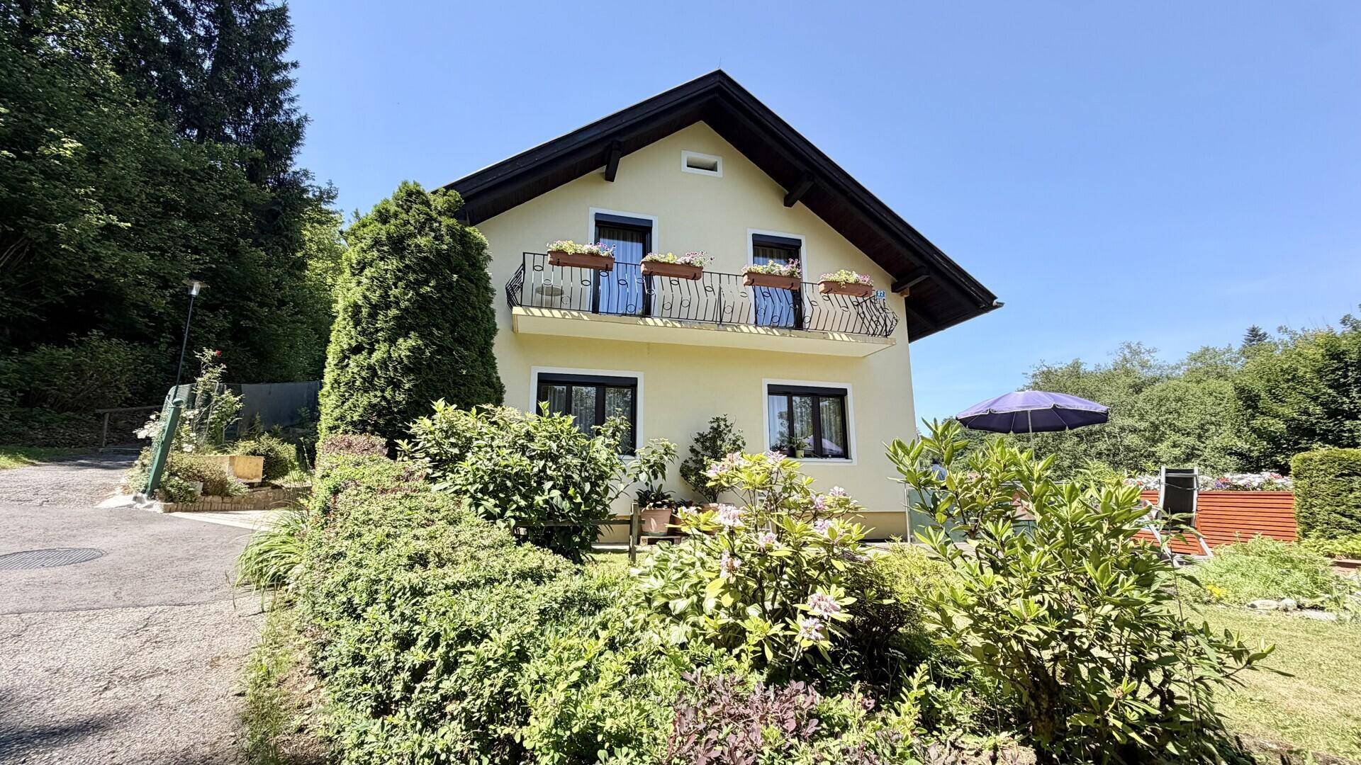 Ansicht des Hauses mit Balkon, gepflegtem Vorgarten und Sonnenschirm im Hintergrund.