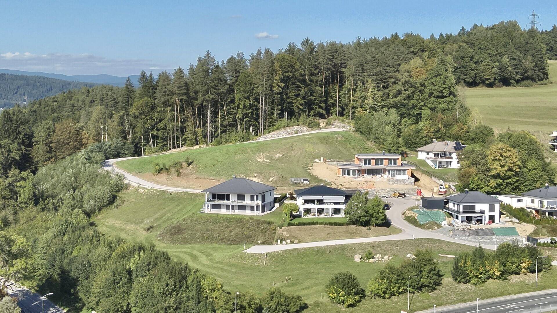 Moderne Häuser auf einem grünen Hügel mit Waldrand und Baustellen in sonniger Umgebung.