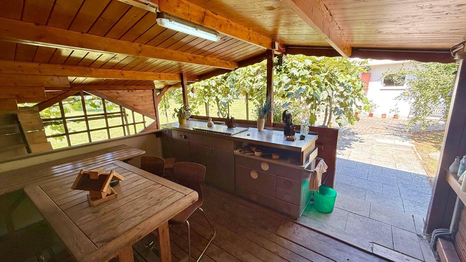 Überdachter Terrassenbereich aus Holz mit Esstisch und Arbeitsfläche, umgeben von Weinreben.