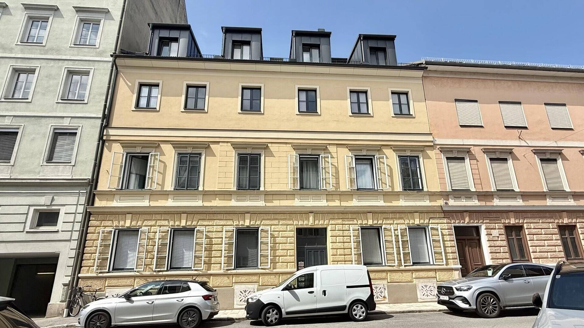 Gelbe Fassade eines Mehrfamilienhauses mit mehreren Fenstern und Dachgauben an einer Stadtstraße.