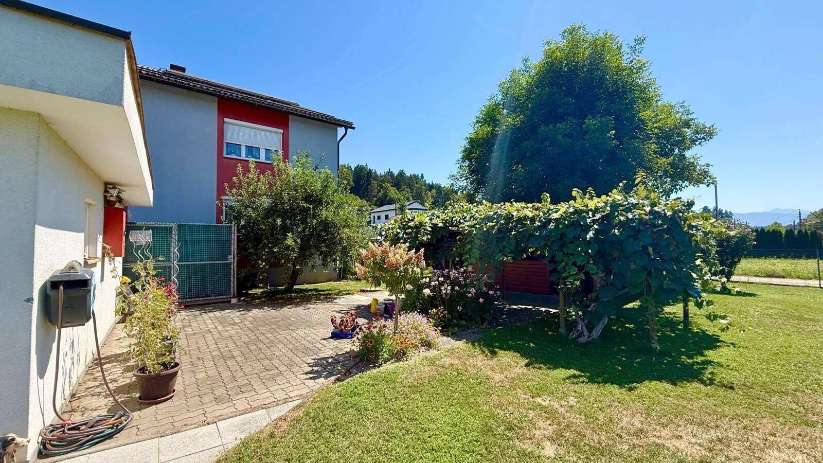 Seitenansicht des Hauses mit gepflastertem Bereich, Blumenbeeten und einer überdachten Laube mit Weinreben.
