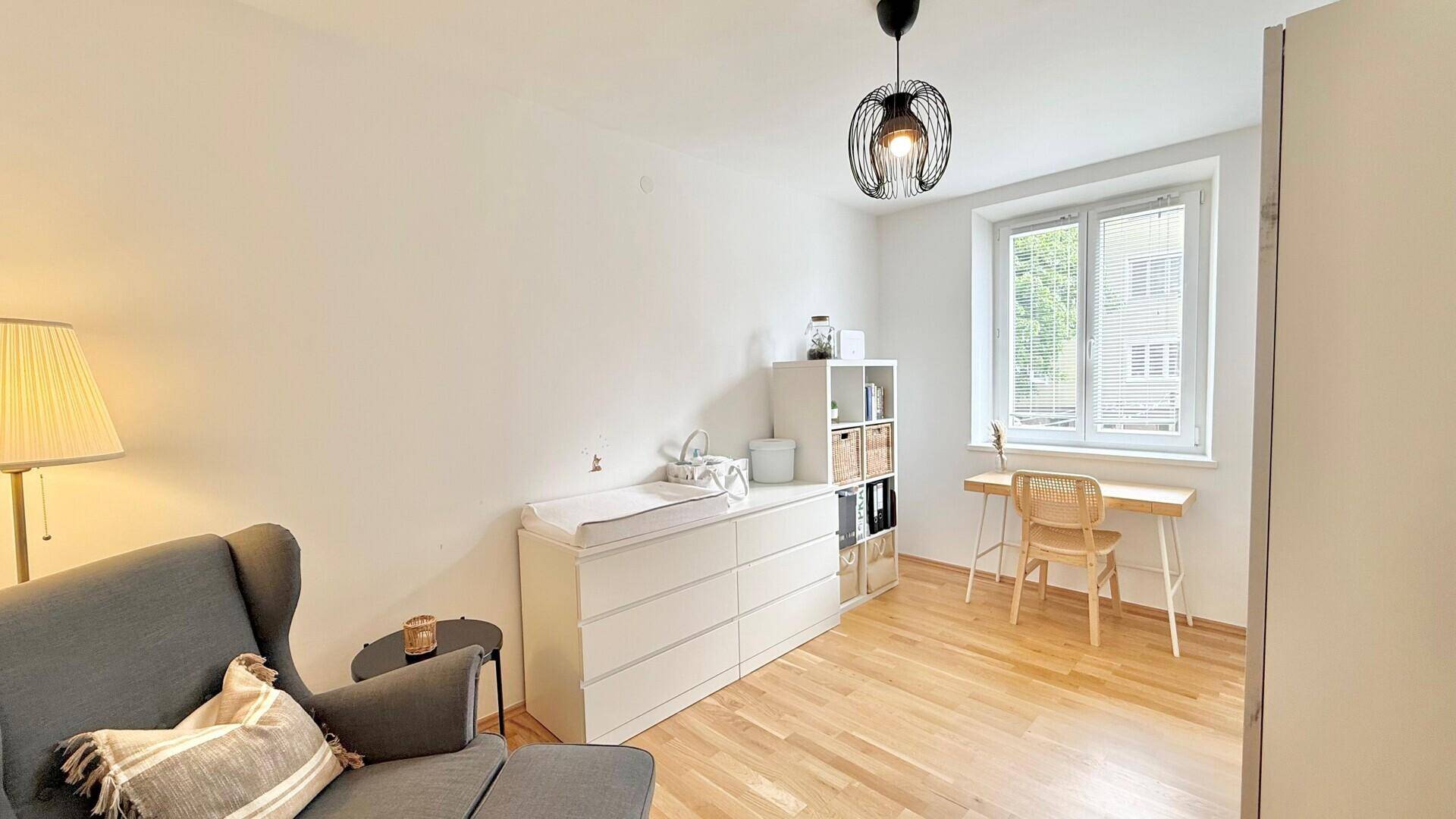 Gemütliches Zimmer mit Wickeltisch, Sessel und Schreibtisch am Fenster.