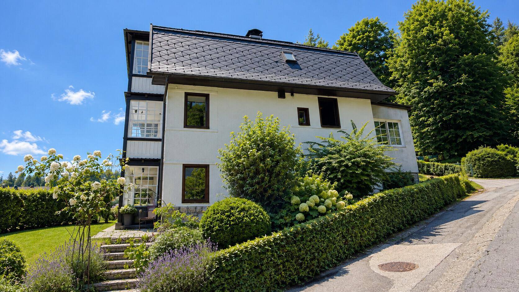 Rückansicht des Hauses mit gepflegtem Garten, Hecken und blühenden Sträuchern unter blauem Himmel.