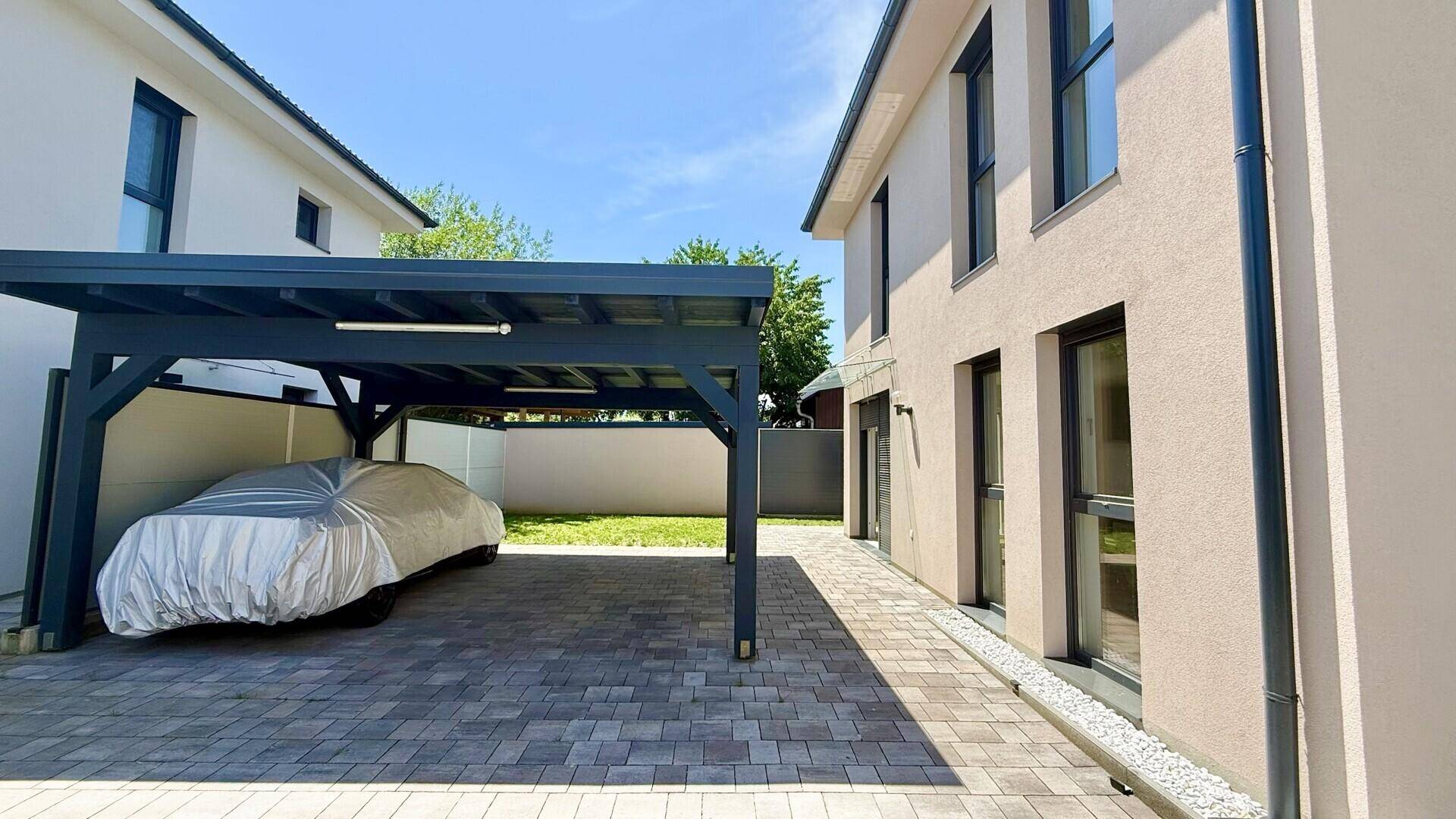 Seitenansicht des modernen Hauses mit Carport und gepflastertem Zugangsbereich unter blauem Himmel.