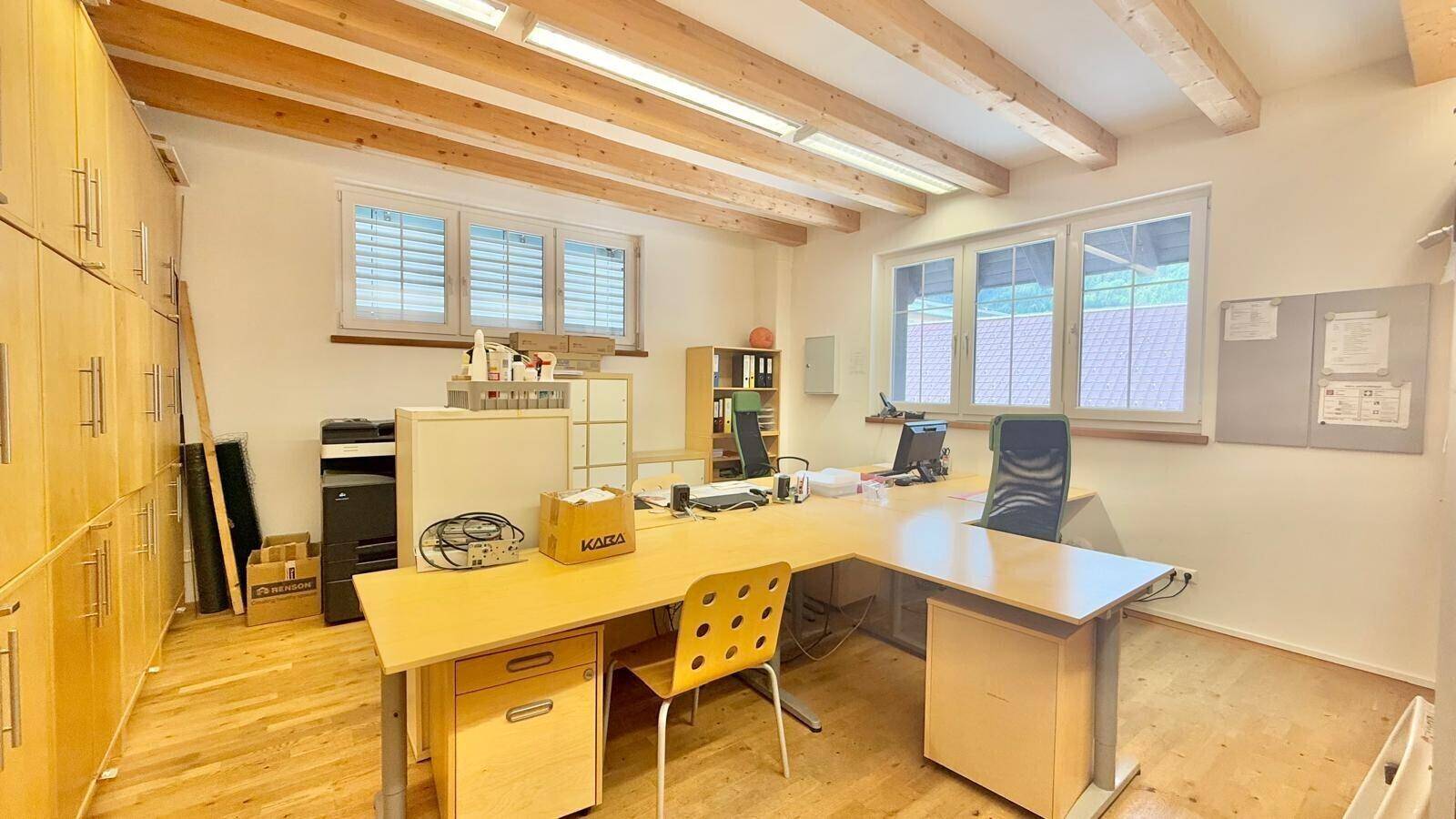 Helles Büro mit Holzbalkendecke, großen Fenstern und mehreren Arbeitsplätzen, ausgestattet mit Holzmöbeln.