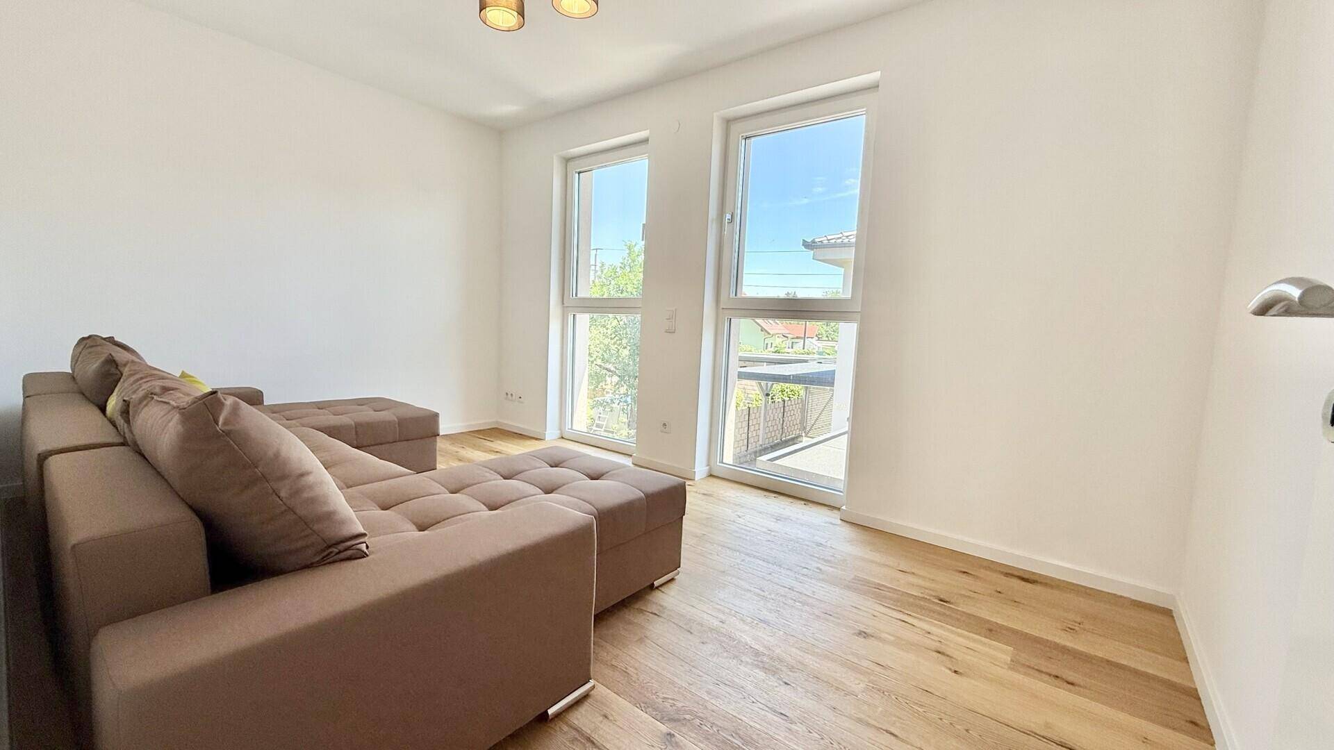 Heller Raum mit Holzboden, großem Sofa und zwei Fenstern, ideal als Wohn- oder Gästezimmer.