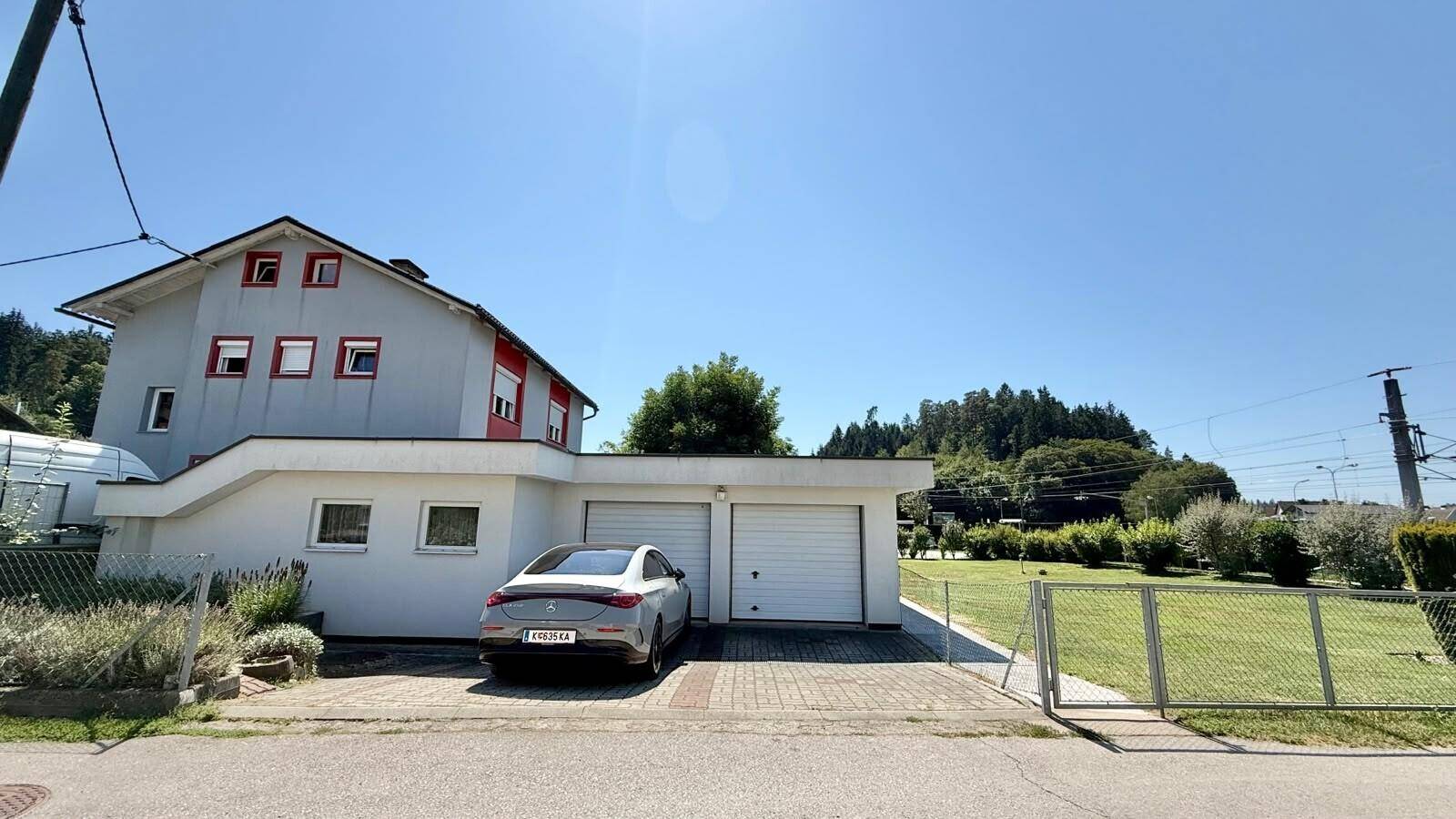 Hausfassade mit Doppelgarage und gepflasterter Einfahrt, umgeben von Grünflächen und Bäumen.