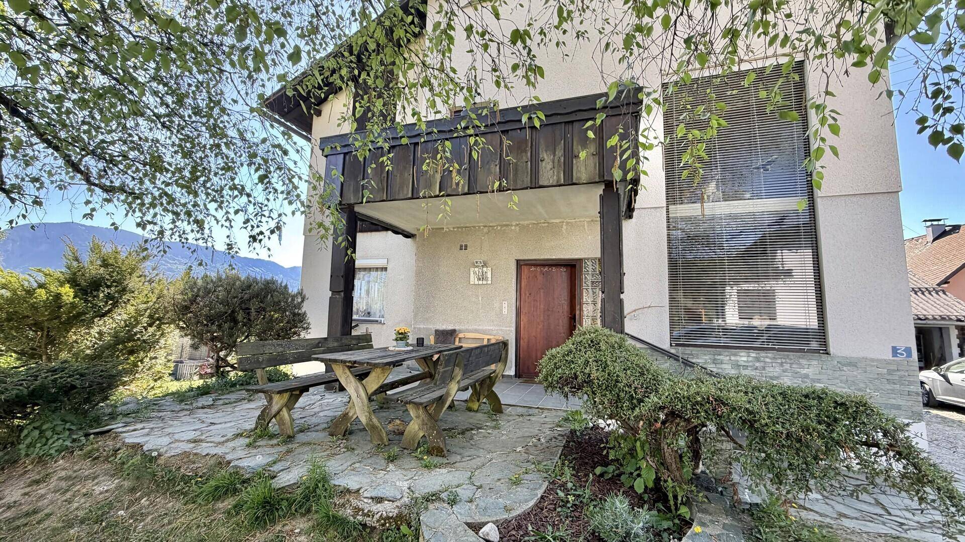 Eingangsbereich des Hauses mit gepflasterter Terrasse und rustikalen Gartenmöbeln.