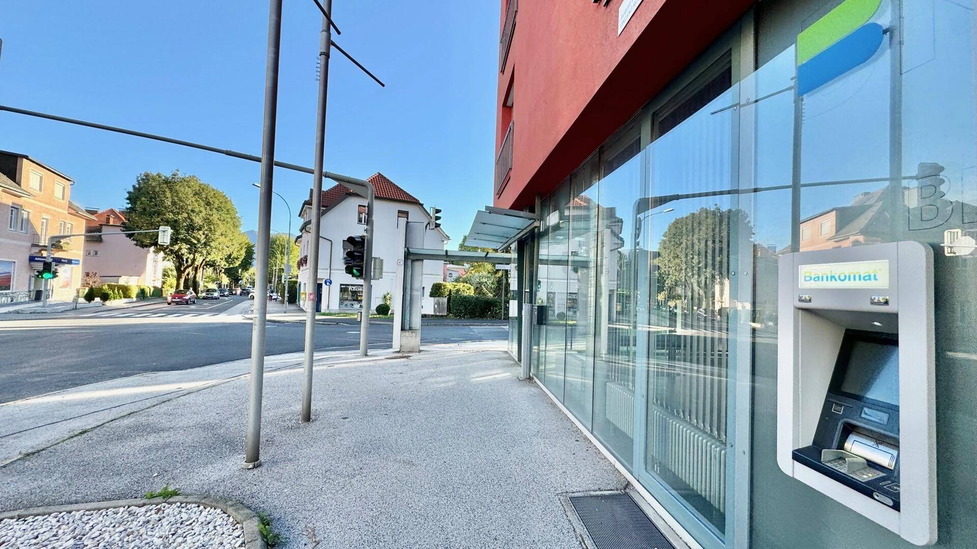 Blick auf die Gebäudefassade mit großen Glasfenstern und einem Bankautomaten an einem sonnigen Tag.