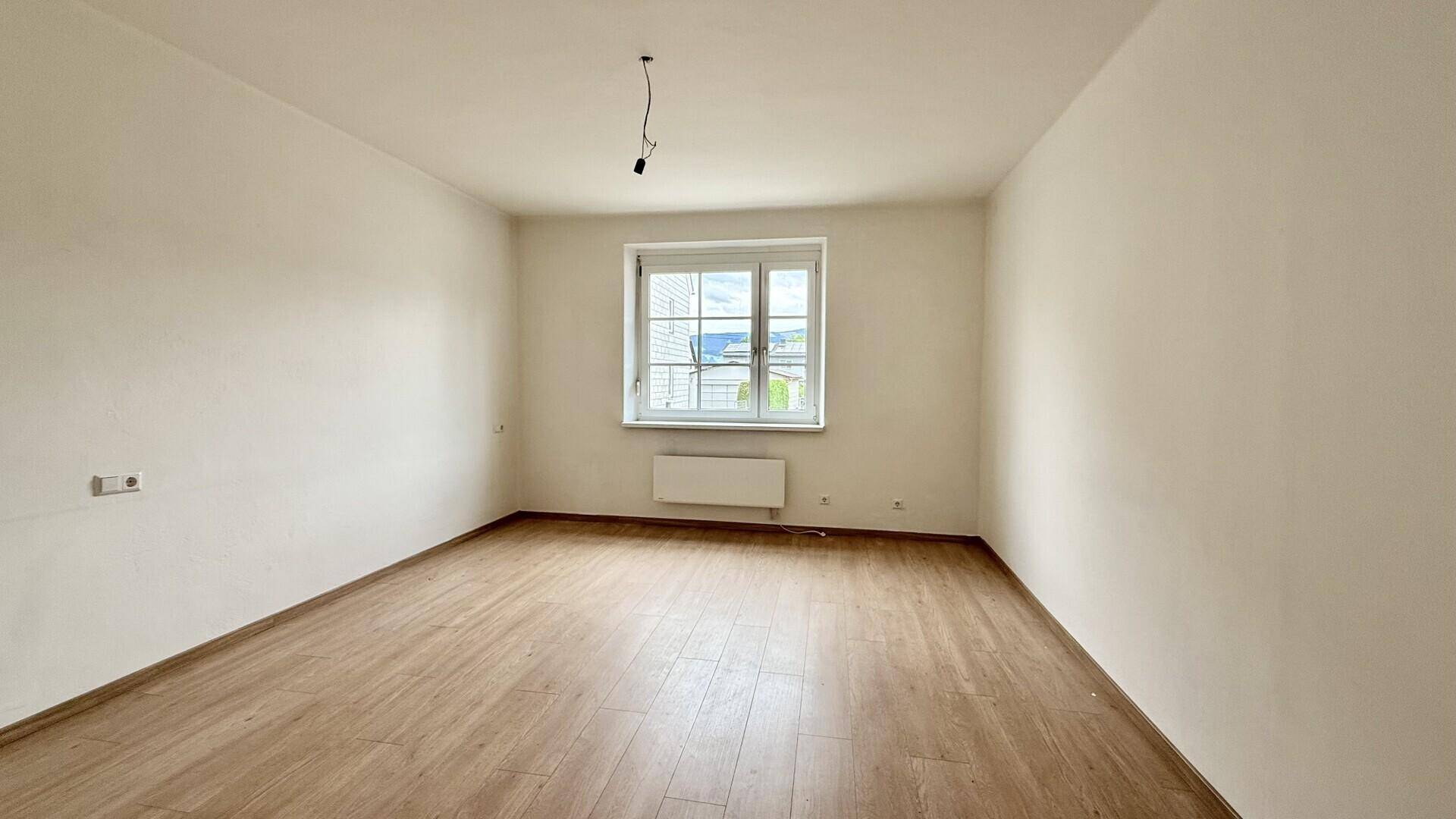Leeres Zimmer mit Laminatboden, hellen Wänden und einem großen Fenster.