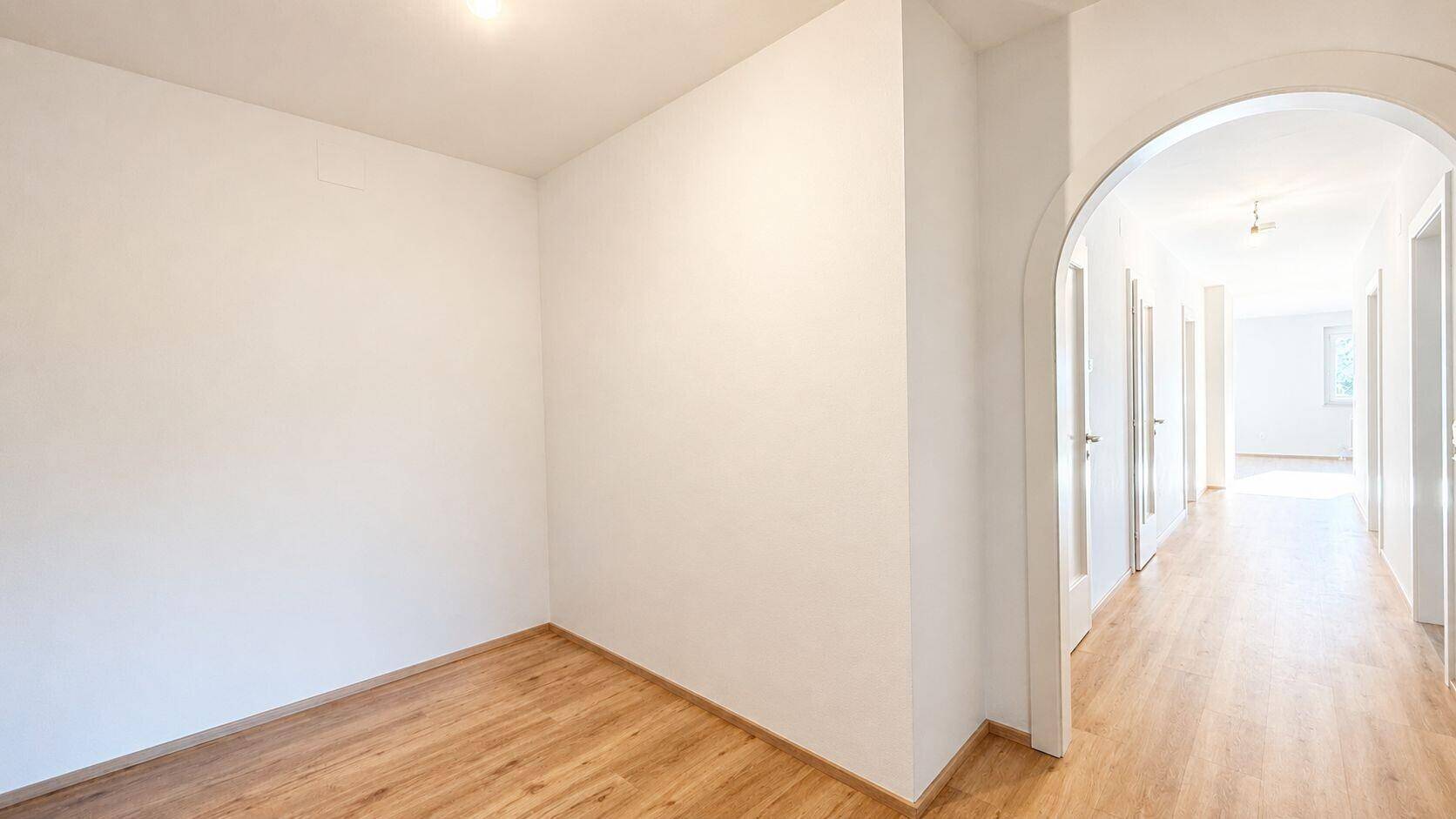 Leerer Raum mit hellem Laminatboden und einem Rundbogen zum Flur.