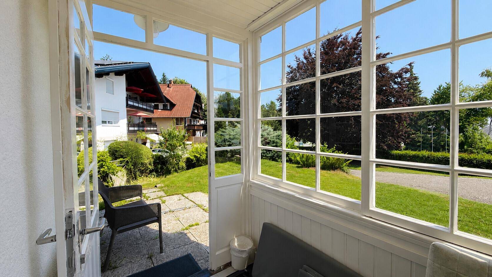 Heller Wintergarten mit großen Fenstern und Blick auf den Garten und umliegende Gebäude.