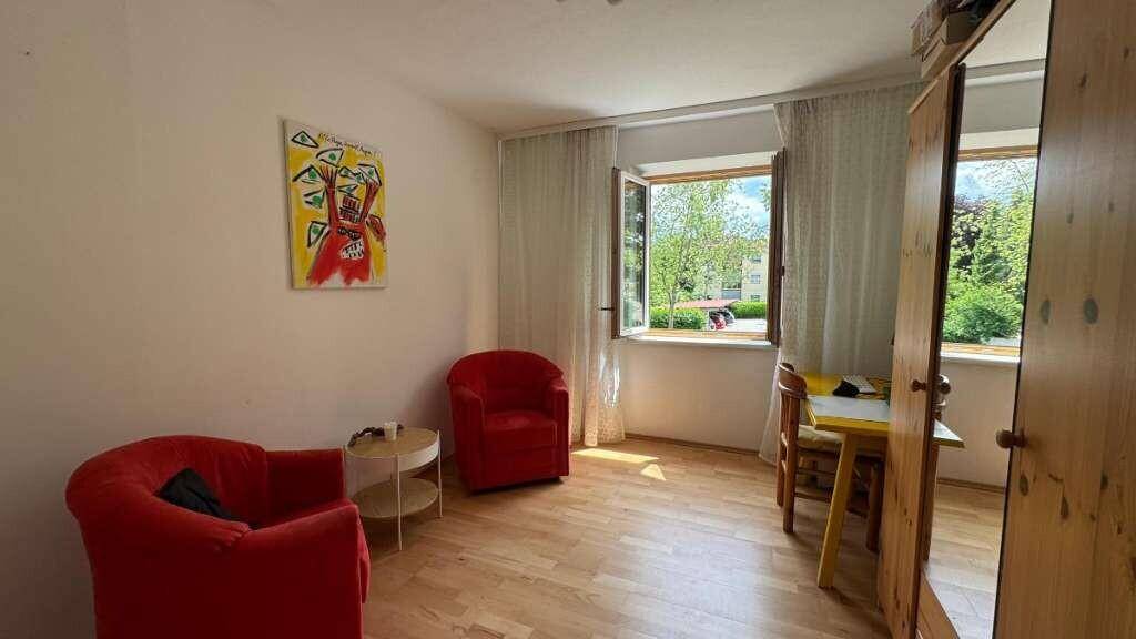 Helles Zimmer mit zwei roten Sesseln, Holzboden und Fenster mit Blick ins Freie.