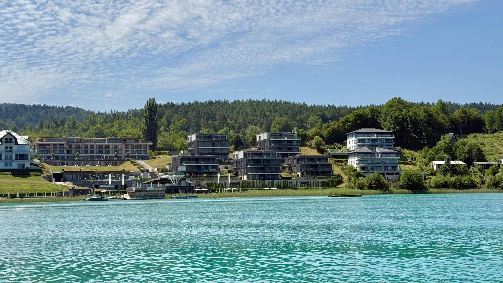 Weitläufiger Blick auf die moderne Wohnanlage am Seeufer vor einer bewaldeten Hügellandschaft.