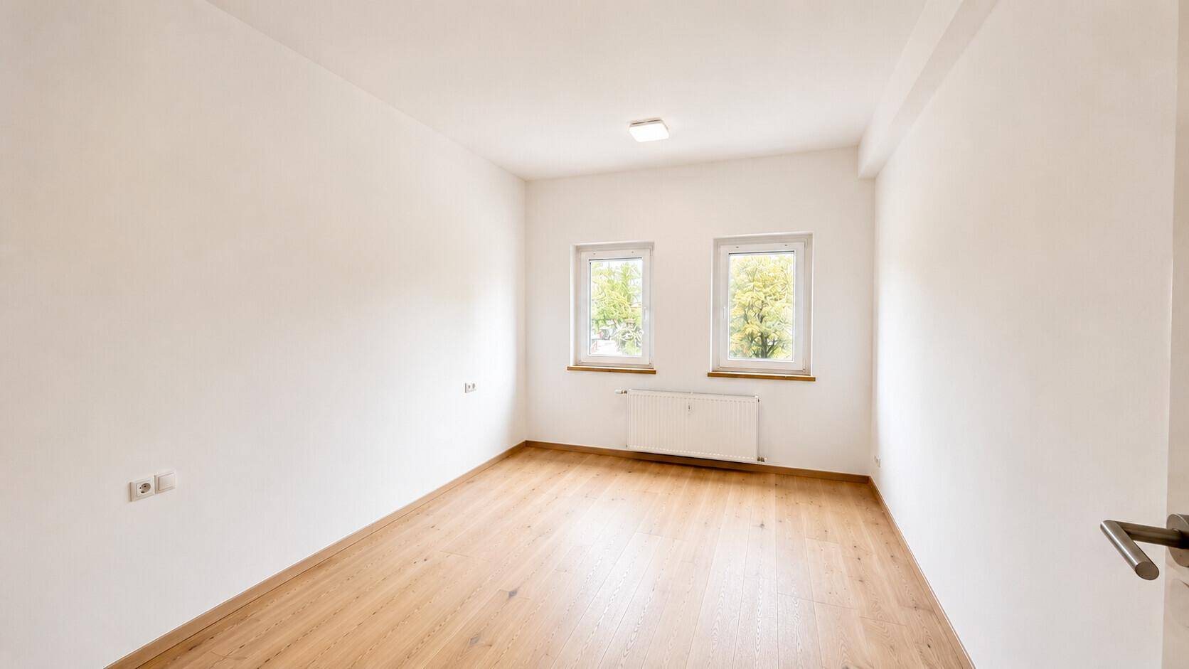 Helles Zimmer mit Holzboden und zwei Fenstern, die einen angenehmen Blick ins Grüne bieten.