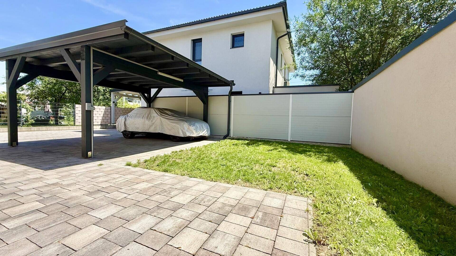 Blick auf den Carport und den kleinen Rasenbereich neben dem modernen Haus unter blauem Himmel.