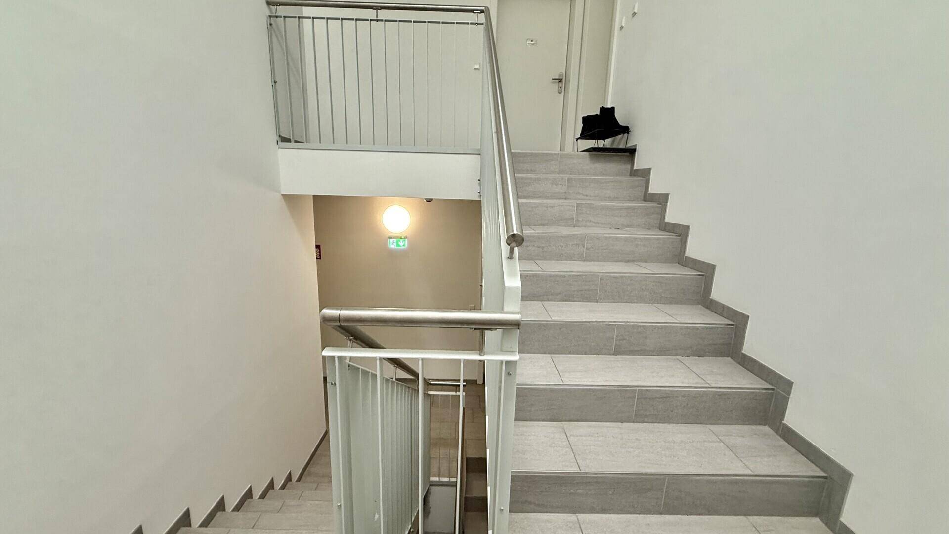Treppenhaus mit Fliesenboden und Edelstahlgeländer, das die verschiedenen Etagen des Gebäudes verbindet.