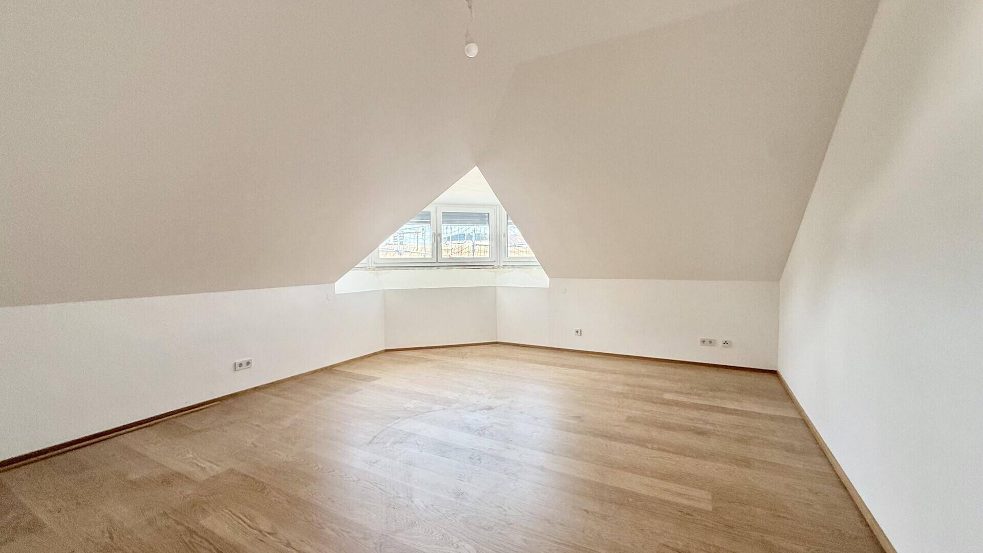 Geräumiger, heller Raum im Dachgeschoss mit Holzboden und schrägen Wänden.