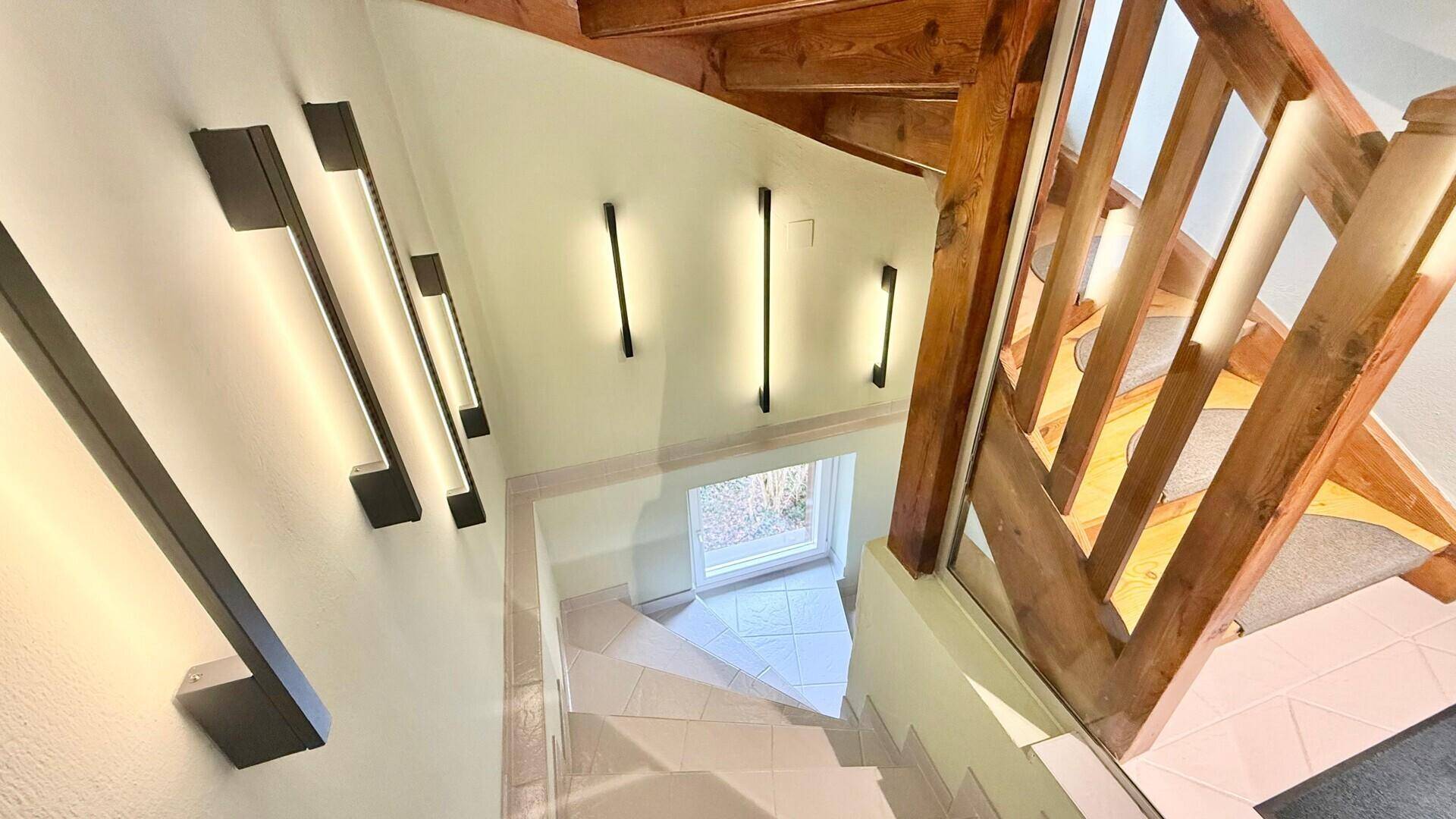 Blick von oben auf die Holztreppe mit moderner Wandbeleuchtung und hellem Fliesenboden.