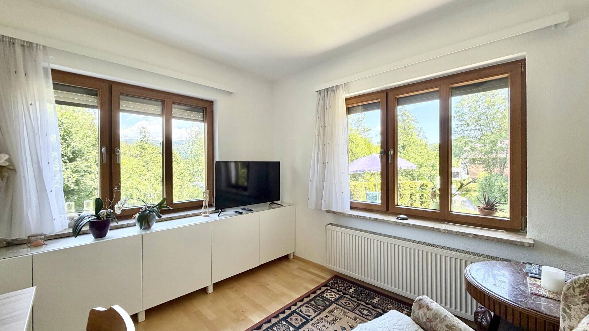Heller Wohnbereich mit großen Fenstern, die viel Tageslicht hereinlassen und eine grüne Aussicht bieten.
