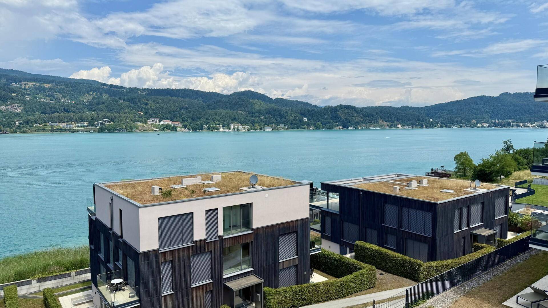 Blick auf moderne Gebäude mit Gründächern, den See und die umliegende Berglandschaft.