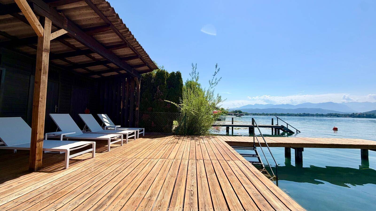 Weitläufige Holzterrasse mit mehreren Liegestühlen und Zugang zu einem langen Holzsteg am See.