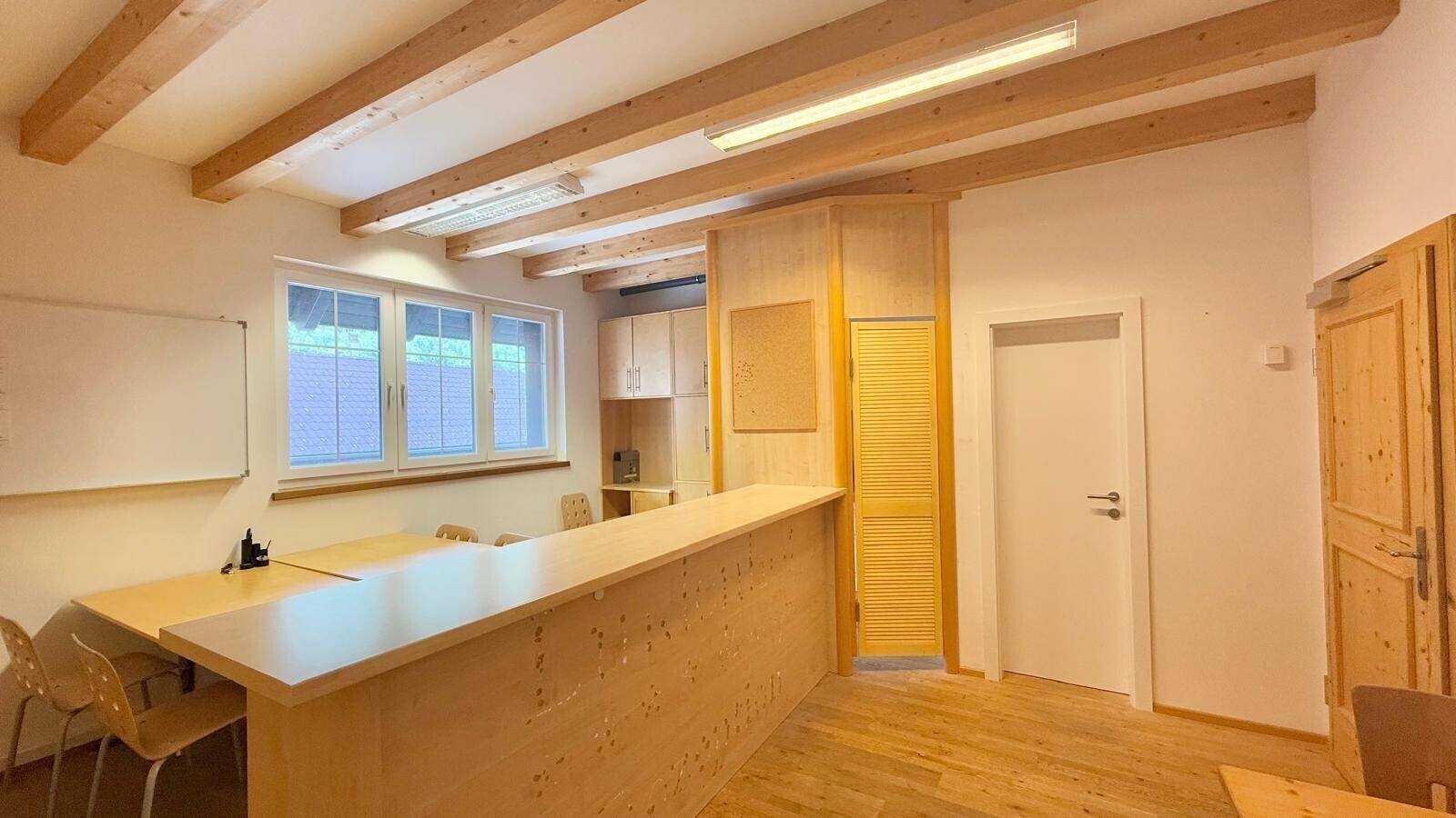 Heller Bürobereich mit Holzbalkendecke, großen Fenstern und einer langen Holztheke, ideal für Empfang oder Besprechungen.