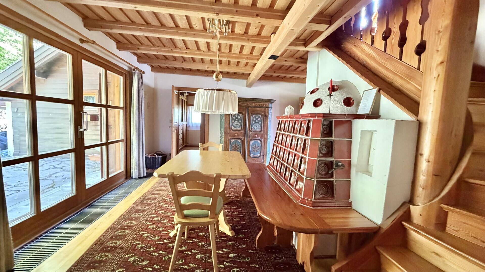 Traditioneller Essbereich mit Holztisch, Kachelofen und offener Holztreppe zum Obergeschoss.