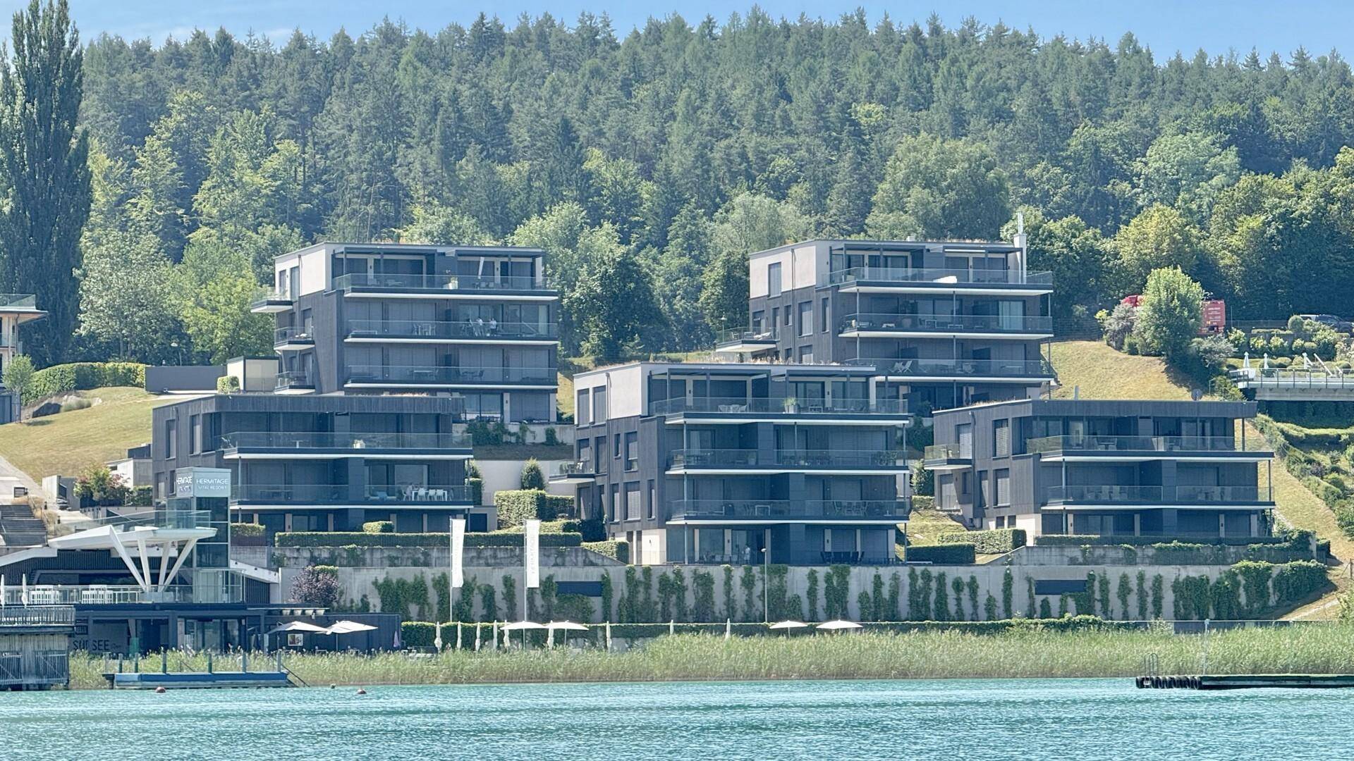 Moderne Wohnanlage am Seeufer mit gestuften Gebäuden und üppiger Vegetation im Hintergrund.