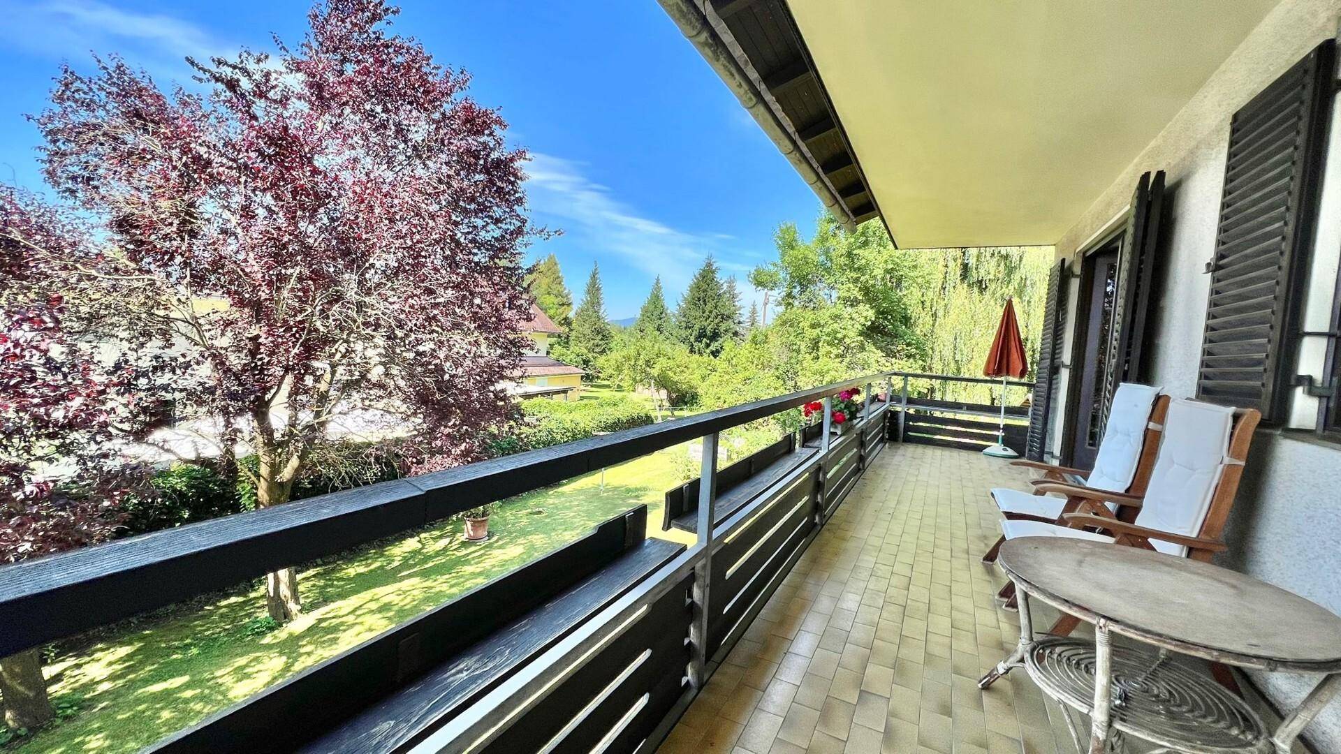 Langer Balkon mit Fliesenboden und Sitzgelegenheiten, umgeben von Bäumen und Natur.