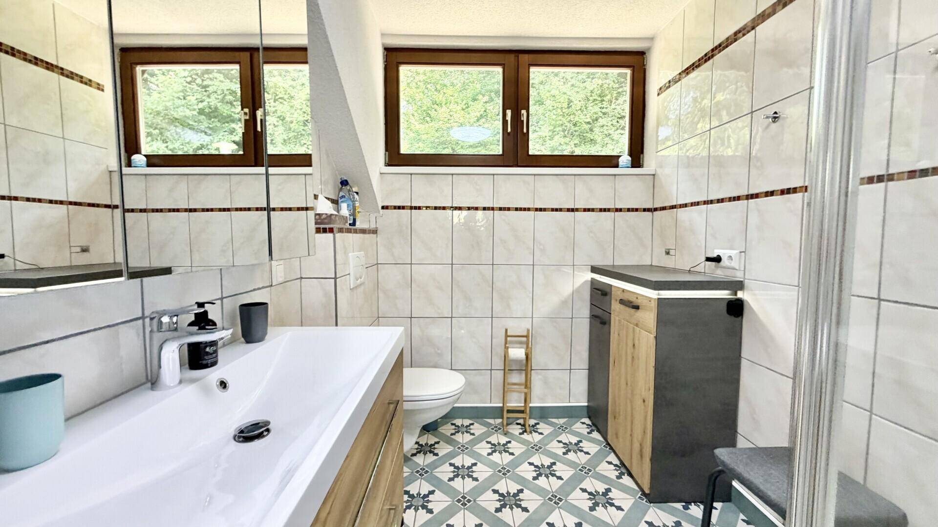 Modern gefliestes Badezimmer mit Duschbereich, Waschbecken und zwei Fenstern für gute Belüftung.