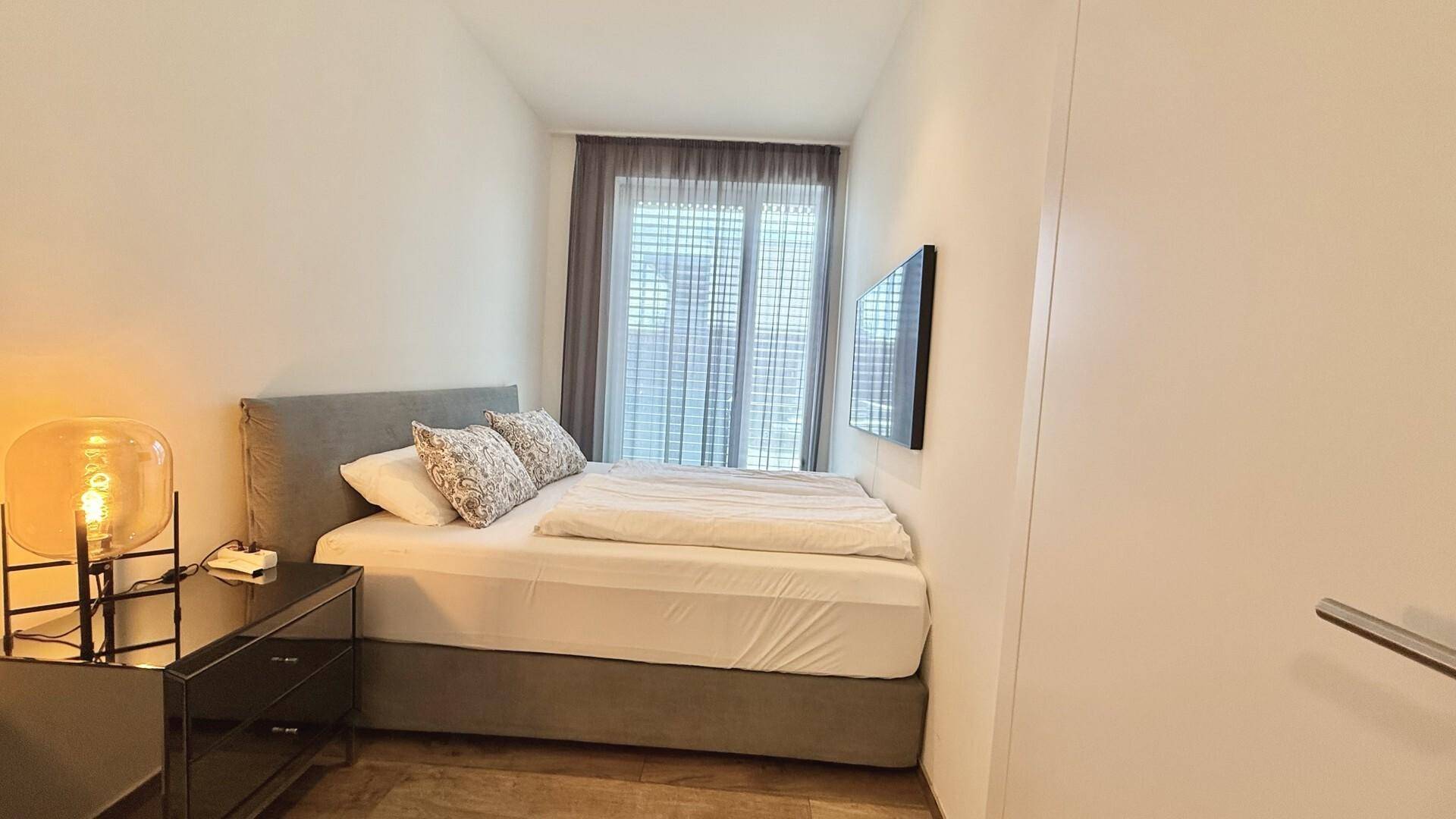 Kleines Schlafzimmer mit Doppelbett, Nachttischlampe und wandmontiertem Fernseher.