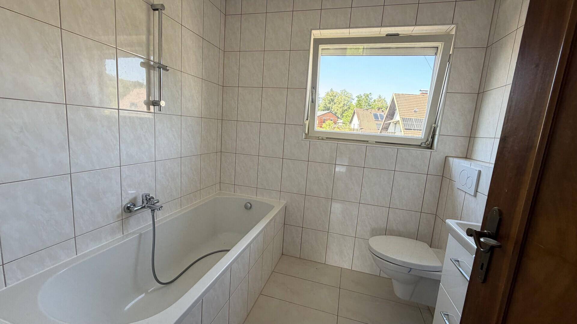 Badezimmer mit Badewanne, WC und Fenster, komplett gefliest.