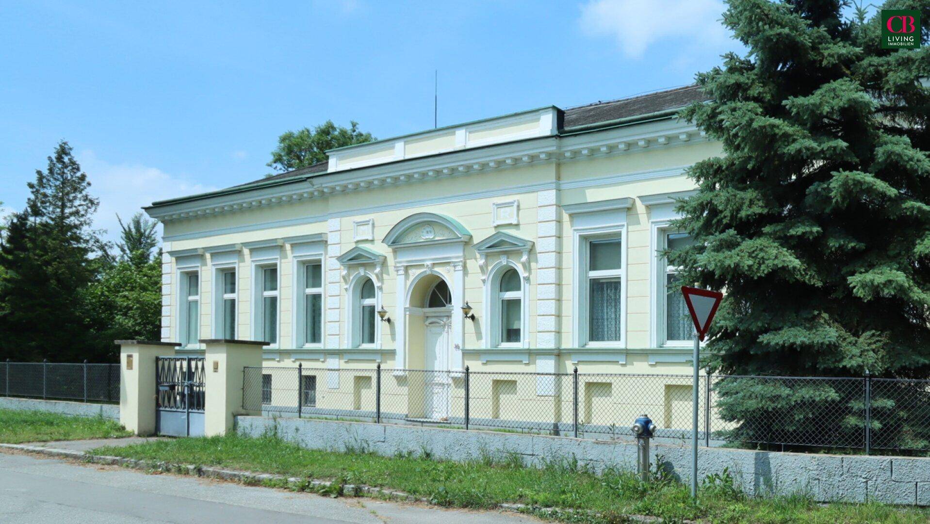 Klassische Fassade eines historischen Gebäudes, umgeben von grünen Bäumen an einem sonnigen Tag.