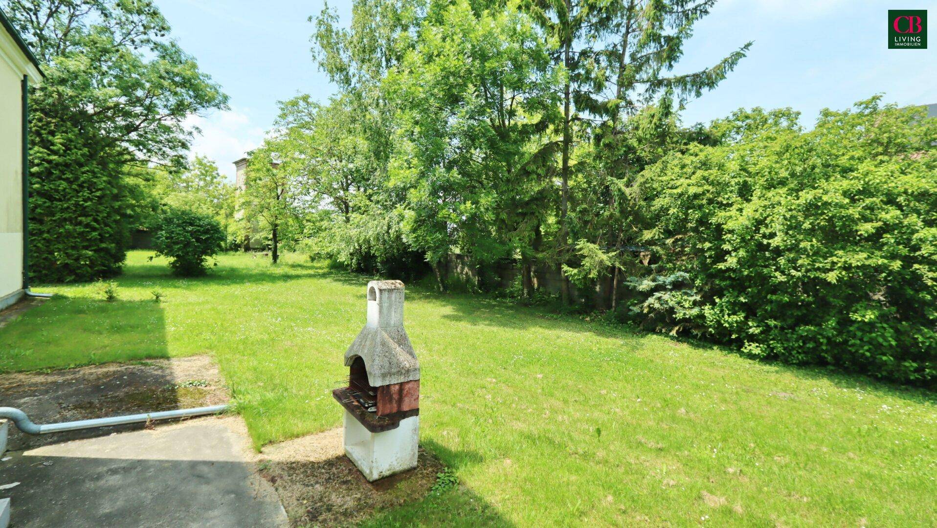 Grüner Garten mit einem gemauerten Grillplatz und hohen Bäumen im Hintergrund.