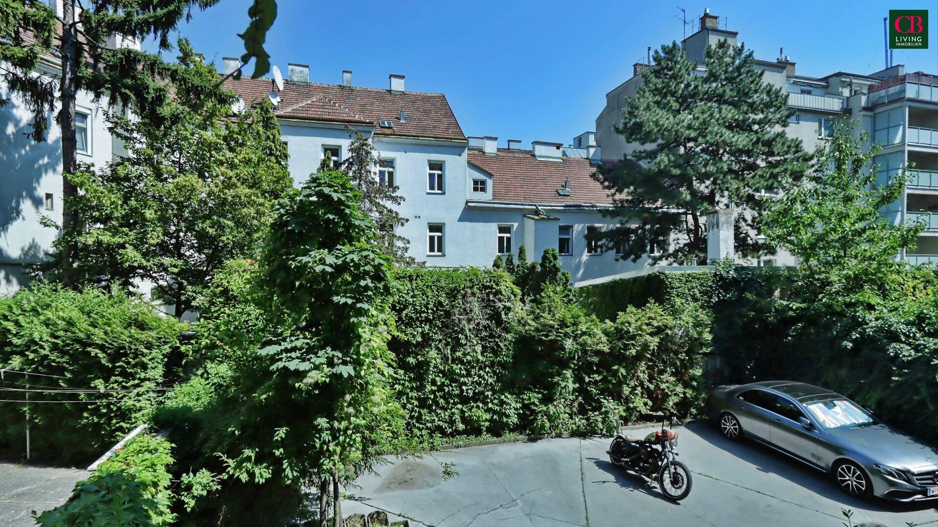 Außenansicht eines Gebäudes mit viel Grün, Bäumen und geparkten Fahrzeugen im Hof.