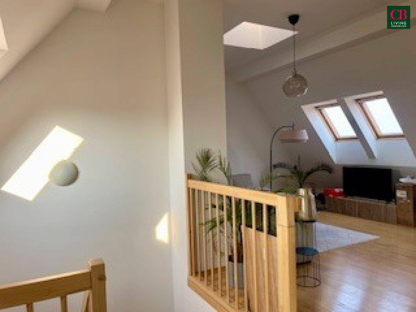 Blick vom Treppenabsatz in das helle Dachgeschoss-Wohnzimmer mit Holzboden und Dachfenstern.