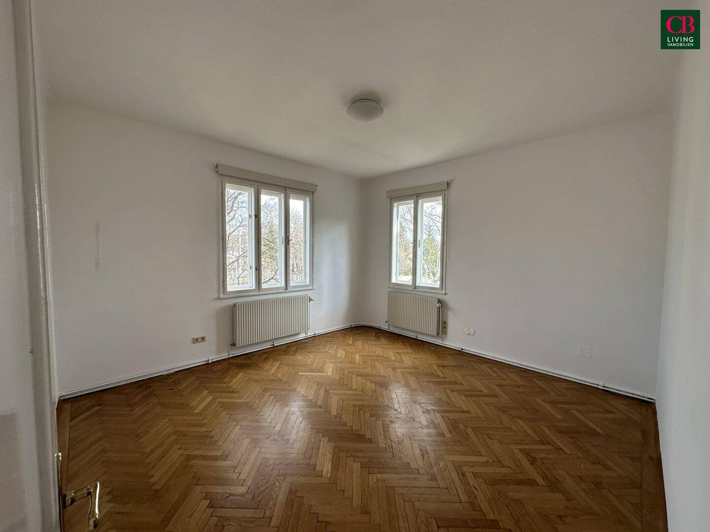 Helles Schlafzimmer mit Fischgrätparkett und zwei Fenstern, unmöbliert.