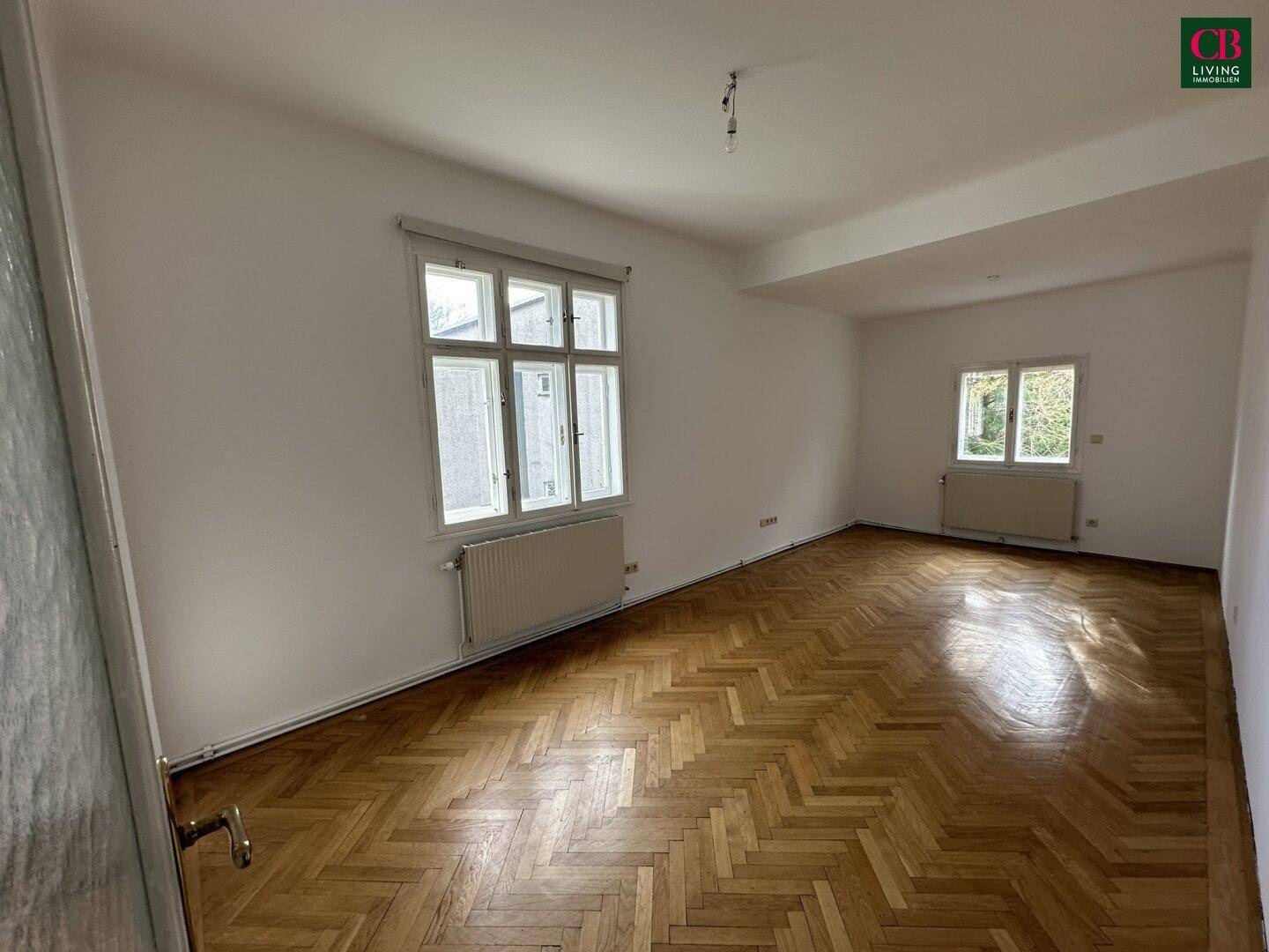 Geräumiges, unmöbliertes Zimmer mit Fischgrätparkett und großen Fenstern.