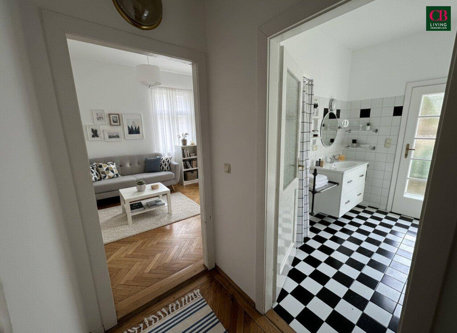 Flur mit Blick in ein möbliertes Wohnzimmer und ein Badezimmer mit Schachbrettmuster-Fliesen.
