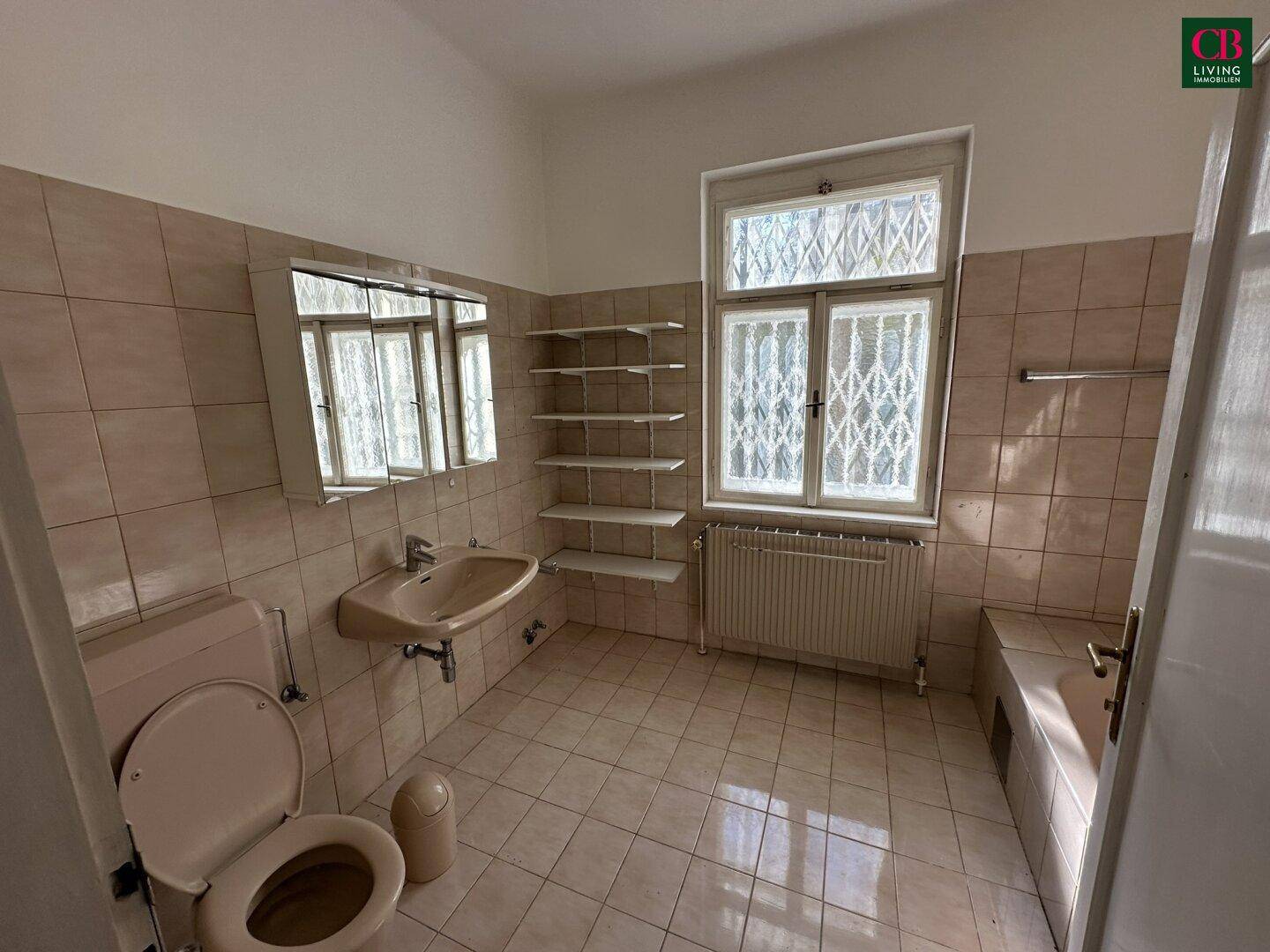 Älteres Badezimmer mit beigefarbenen Fliesen, Badewanne und Fenster.