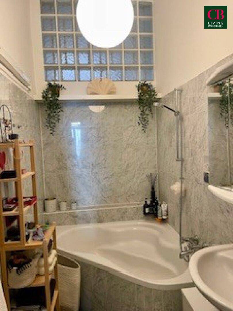Badezimmer mit Eckbadewanne, hellgrauen Fliesen und einem Glasbausteinfenster, das für natürliches Licht sorgt.