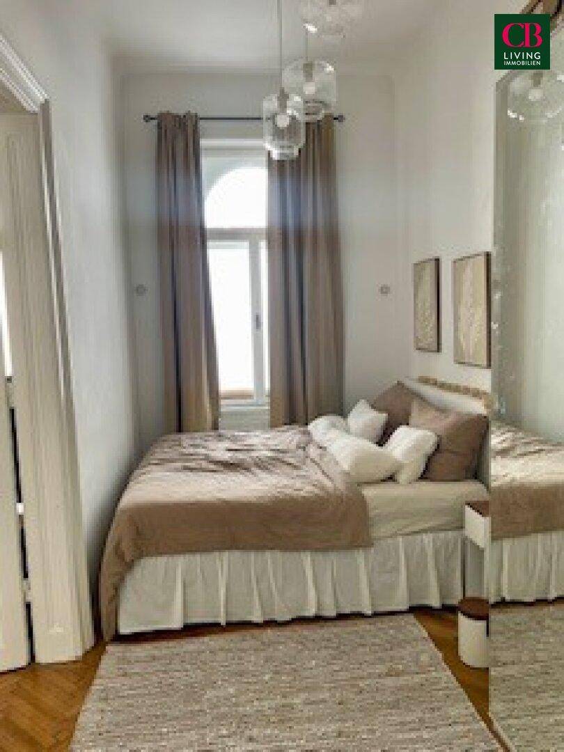 Schlafzimmer mit einem bequemen Bett, einem hohen Fenster und hellen Vorhängen, die eine ruhige Atmosphäre schaffen.