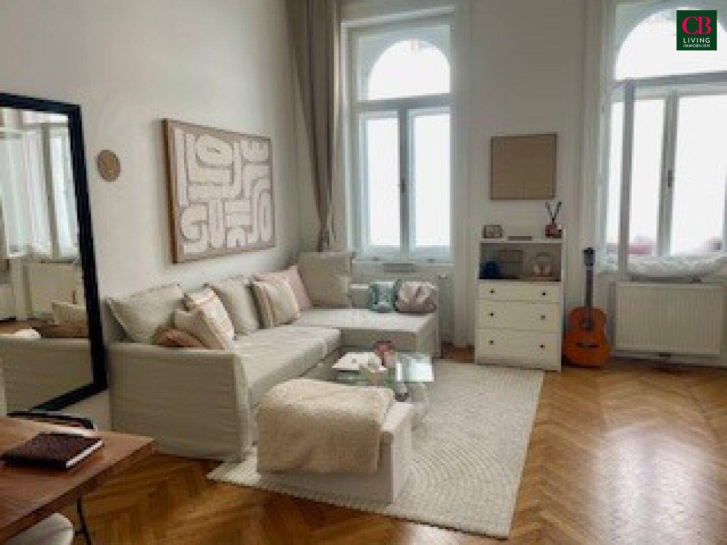 Helles Wohnzimmer mit beigem Ecksofa, Holzboden und hohen Fenstern, die viel Tageslicht hereinlassen.