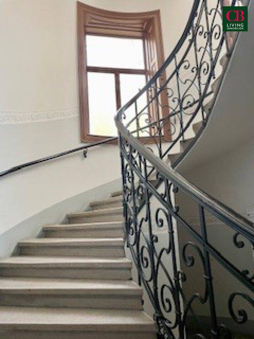 Elegantes Treppenhaus eines Altbaus mit geschwungenem schmiedeeisernem Geländer und hohen Fenstern.