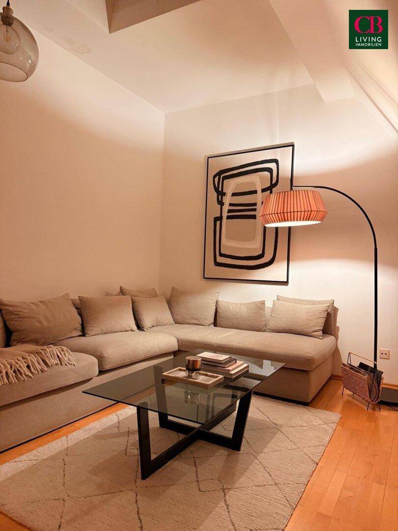 Gemütliches Wohnzimmer mit Ecksofa, Couchtisch aus Glas und Holzboden, beleuchtet von einer Stehlampe.